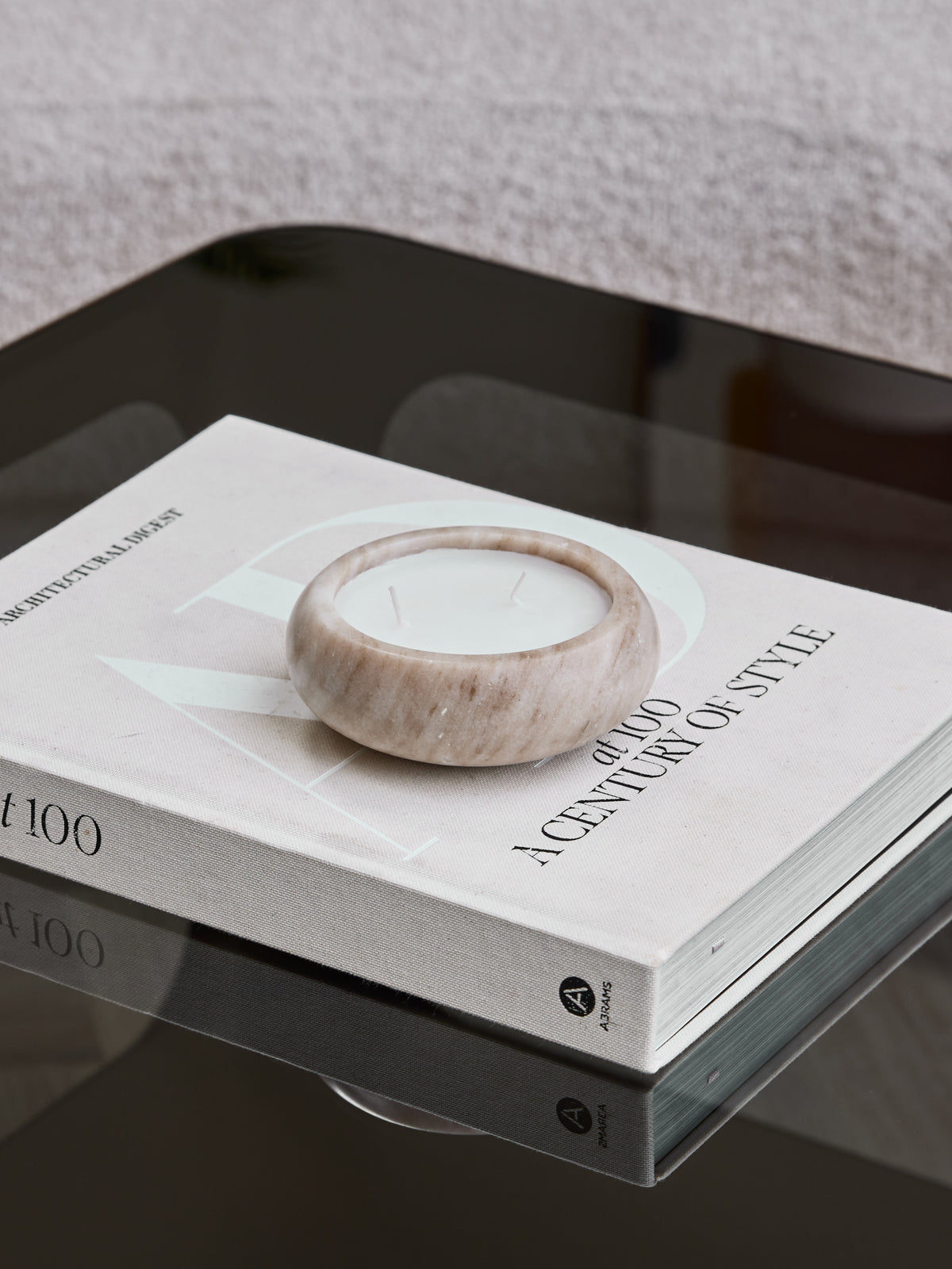 Beige marble candle with two unlit wicks resting on an Architectural Digest book on a reflective dark glass table.