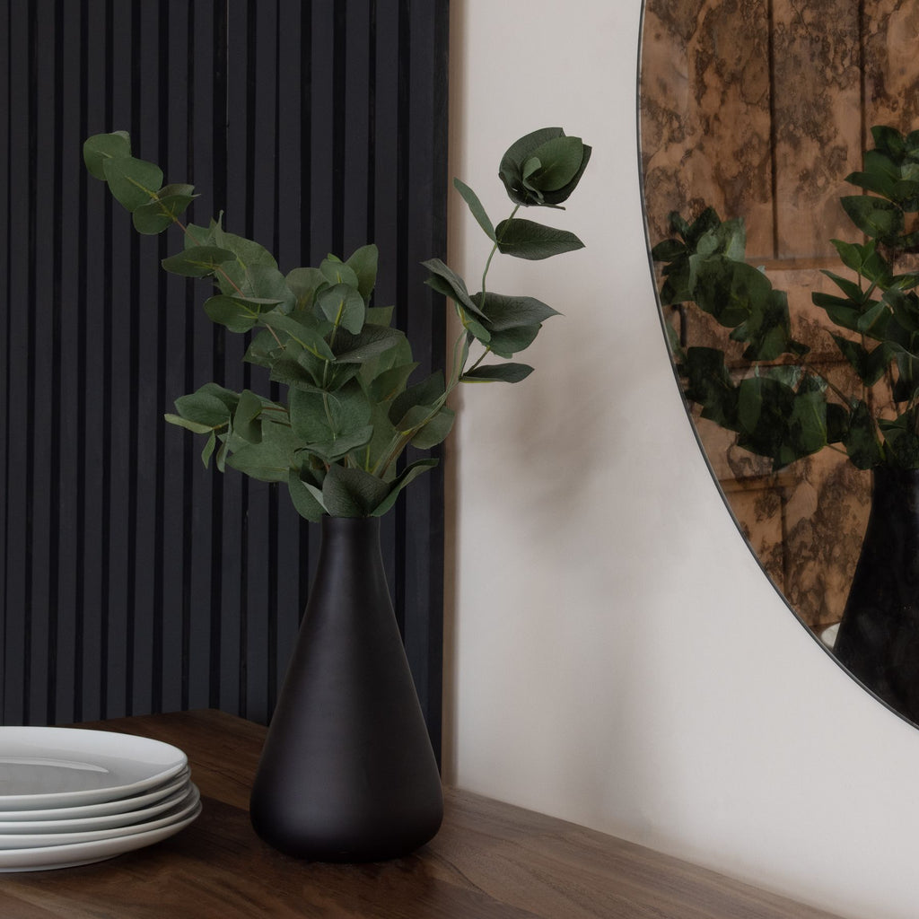 Green artificial foliage arranged in a dark tapered vase on a wooden surface beside stacked white plates, with dark wall panelling and a round mirror behind.