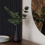 Green artificial foliage arranged in a dark tapered vase on a wooden surface beside stacked white plates, with dark wall panelling and a round mirror behind.