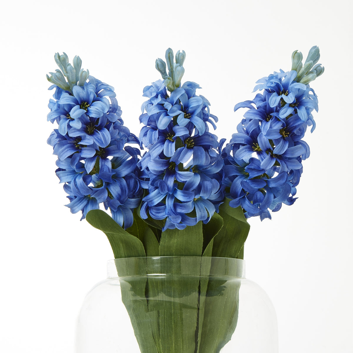 Three blue artificial flower spikes arranged with broad green leaves in a clear rounded vase against white.