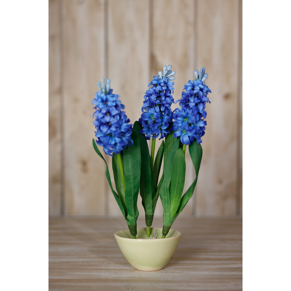 Three blue artificial flower stems displayed in a pale green bowl on a wooden surface against a pale wood-panelled backdrop.