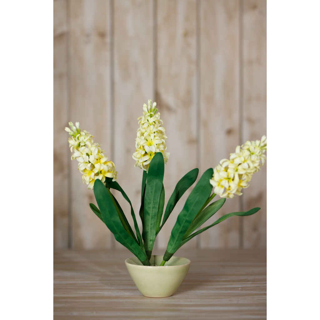 Pale yellow artificial flower stems fanned out in a low pale yellow bowl on a wooden surface in front of timber panelling.