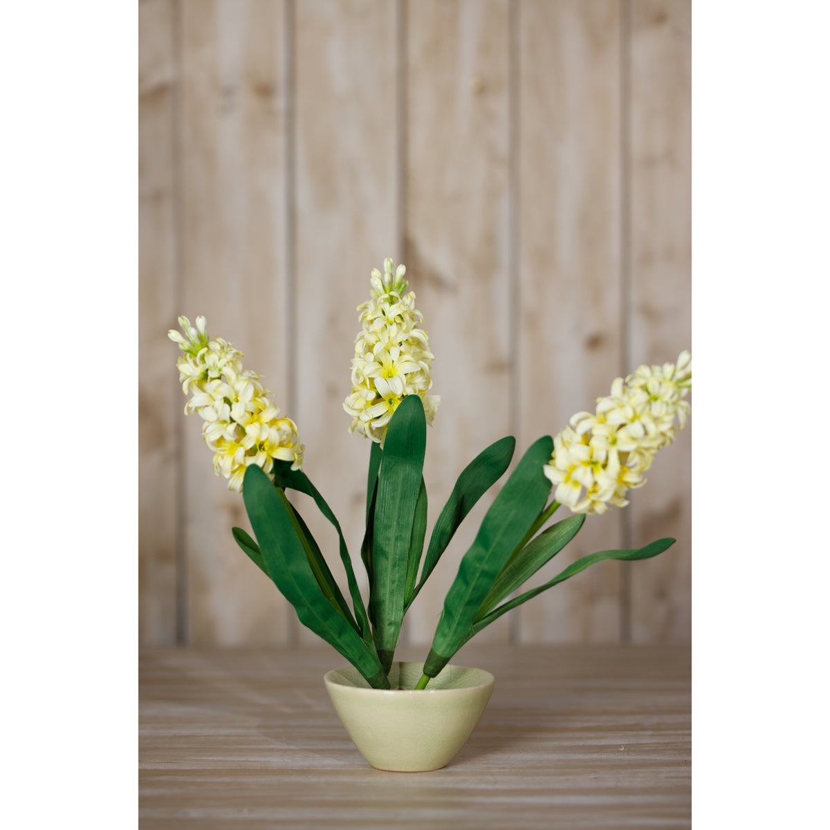 Pale yellow artificial flower stems fanned out in a low pale yellow bowl on a wooden surface in front of timber panelling.