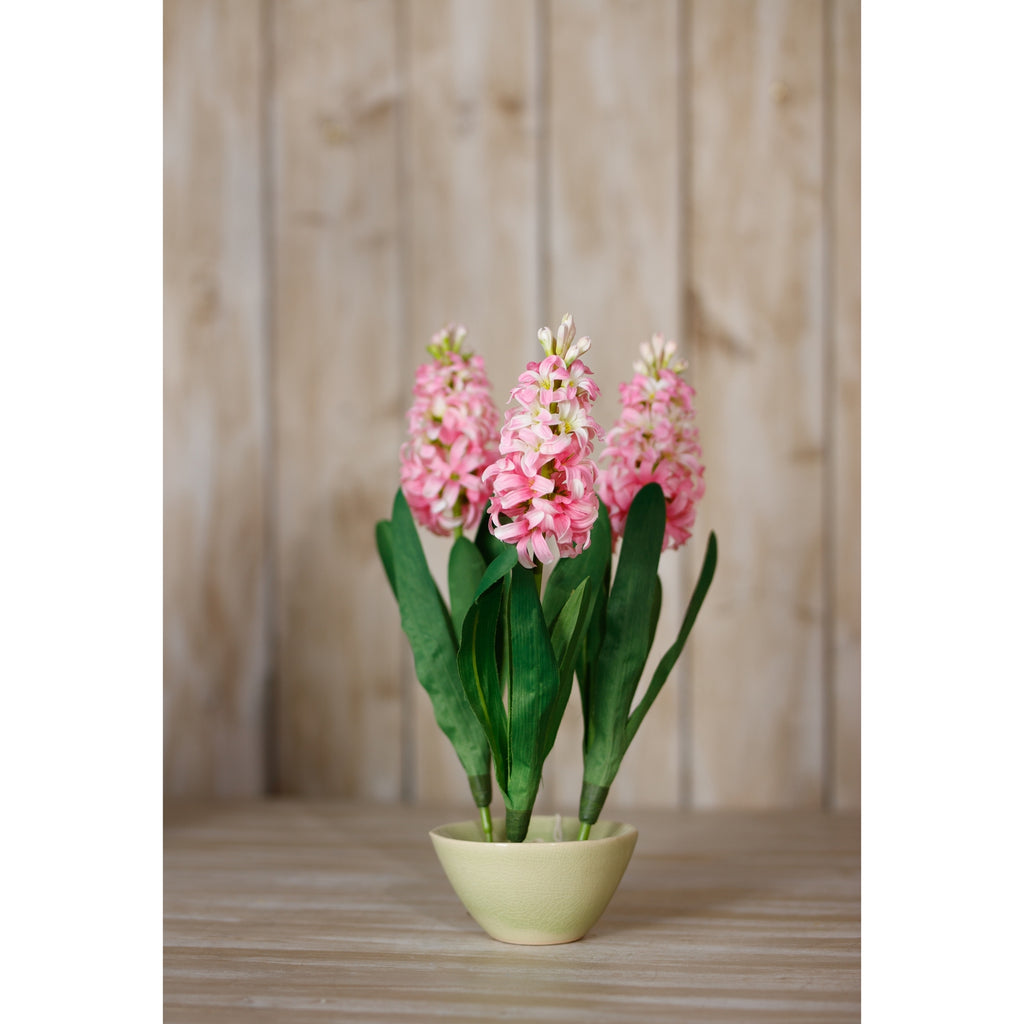 Three pink artificial flower stems arranged in a pale green bowl on a wooden surface against vertical wooden panelling.