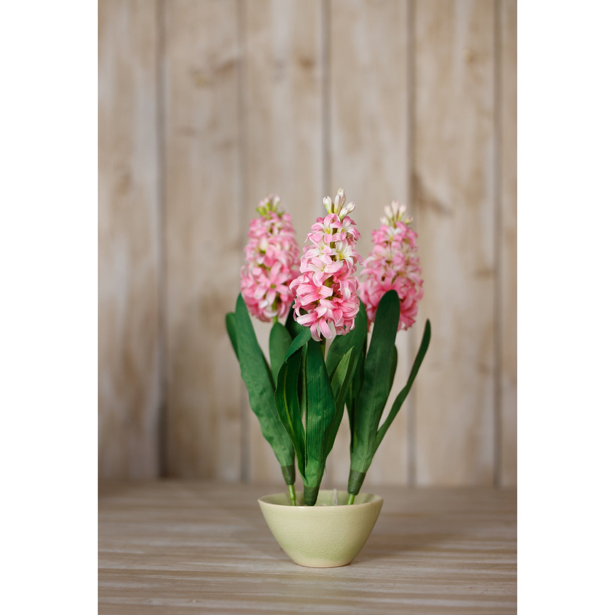 Three pink artificial flower stems arranged in a pale green bowl on a wooden surface against vertical wooden panelling.
