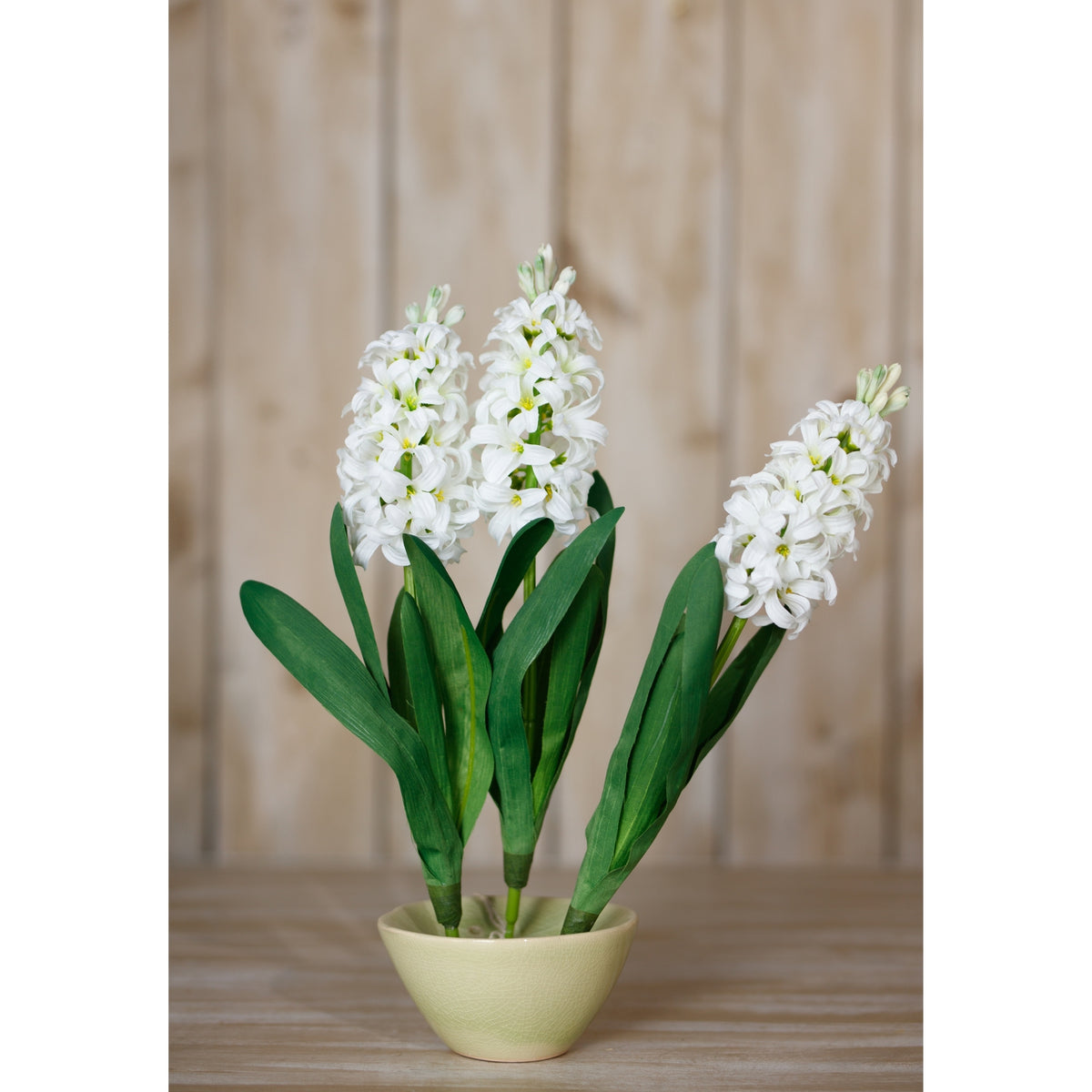 Three white flower spikes and green leaves arranged in a pale green bowl-shaped pot on a wooden surface against vertical wood panels.