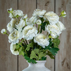 Close view of a multi-stem arrangement of pale artificial flowers and lobed green leaves in a white vase against vertical wood boards.