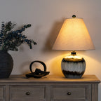 Table lamp shown with a glowing tapered shade on a wooden sideboard beside a dark curved sculpture and a vase of leafy stems.