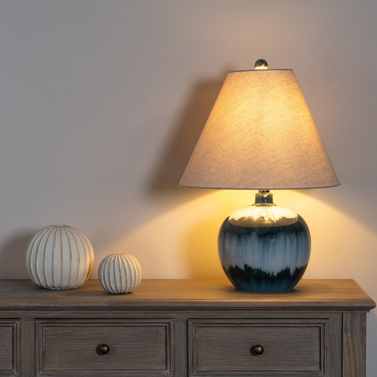 Illuminated blue table lamp on a wooden sideboard beside two pale ribbed ornaments, with warm light visible across the shade and wall