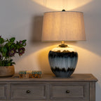 Lamp pictured illuminated on a wooden sideboard beside two stacked books and a pot of dark flowers, with warm light visible on the wall.
