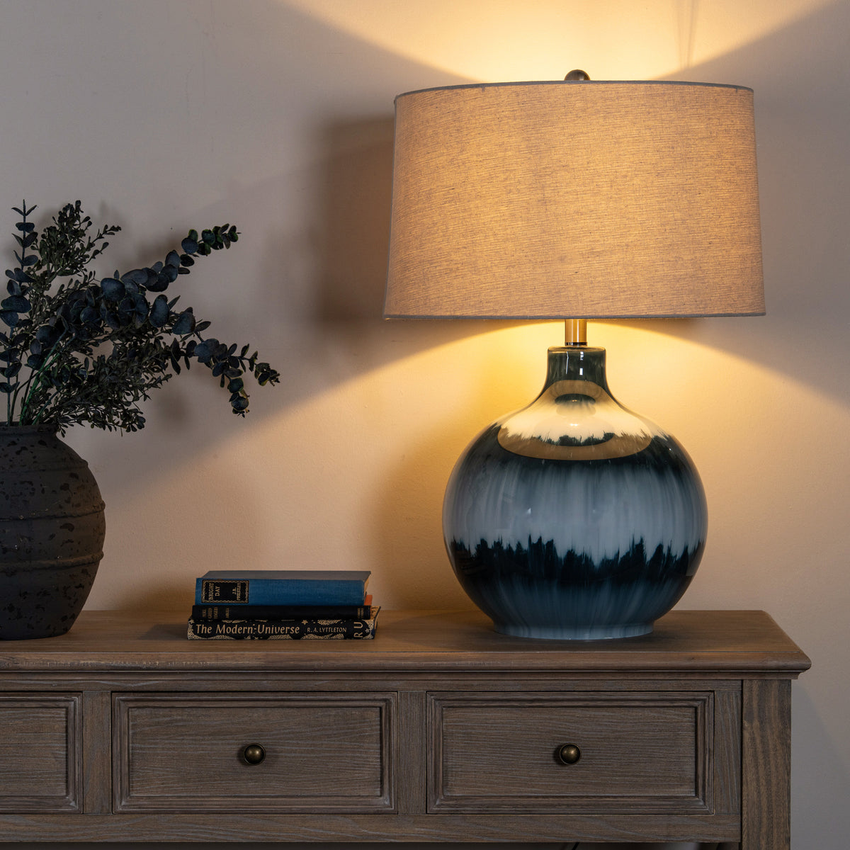 Blue banded table lamp shown illuminated on a wooden sideboard beside stacked books and a vase of leafy stems, with warm light falling across the wall.