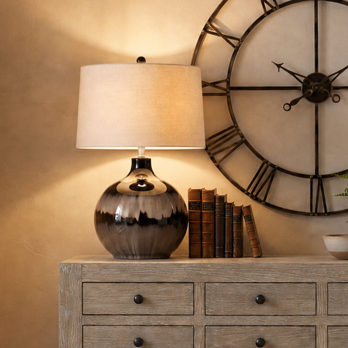 Banded table lamp pictured illuminated on a pale wooden sideboard beside brown books, with a large open-frame wall clock behind.