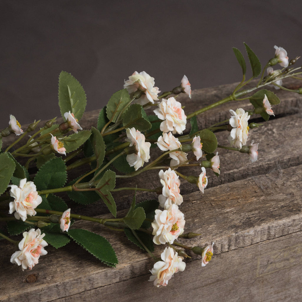 Close-up of artificial meadow rose flowers laid across weathered wood, showing cream petals with pink centres, buds and serrated green leaves.
