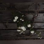 White artificial meadow rose spray laid diagonally across weathered wooden slats among twisting bare branches.