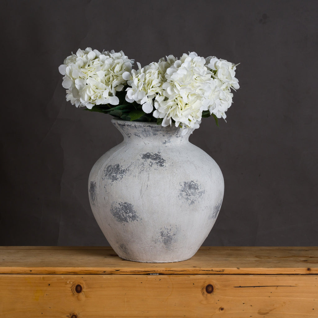 White and grey rounded vase styled with white hydrangea blooms on a wooden sideboard against a dark wall.