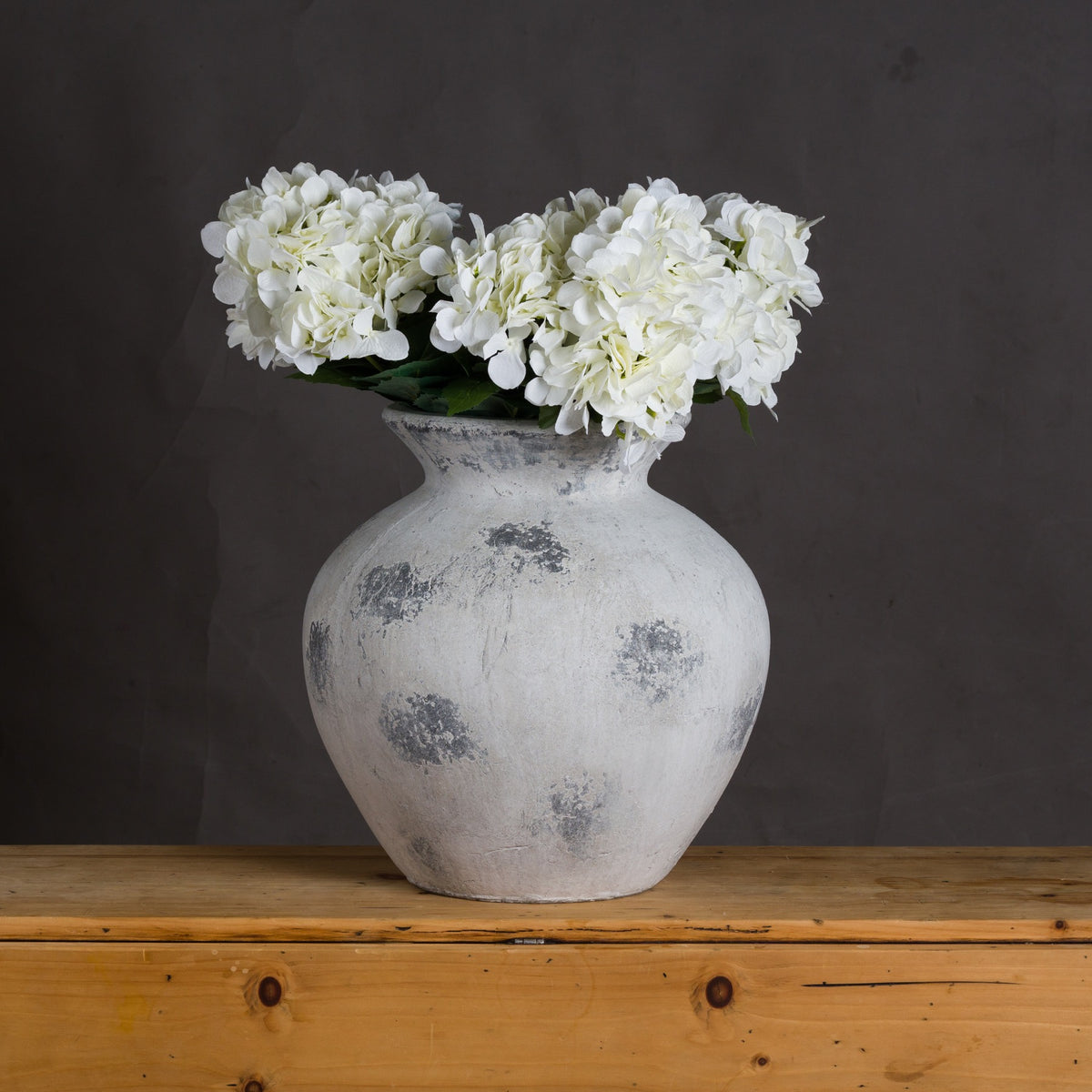 White and grey rounded vase styled with white hydrangea blooms on a wooden sideboard against a dark wall.