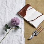 Light-purple artificial flower stem laid diagonally across white cloth beside an open brown-covered notebook and secateurs on a tan surface.