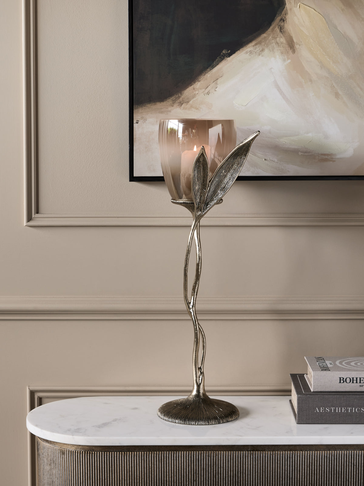 Leaf Stem Hurricane holding a lit candle on a pale console beside stacked books, beneath an abstract painting.