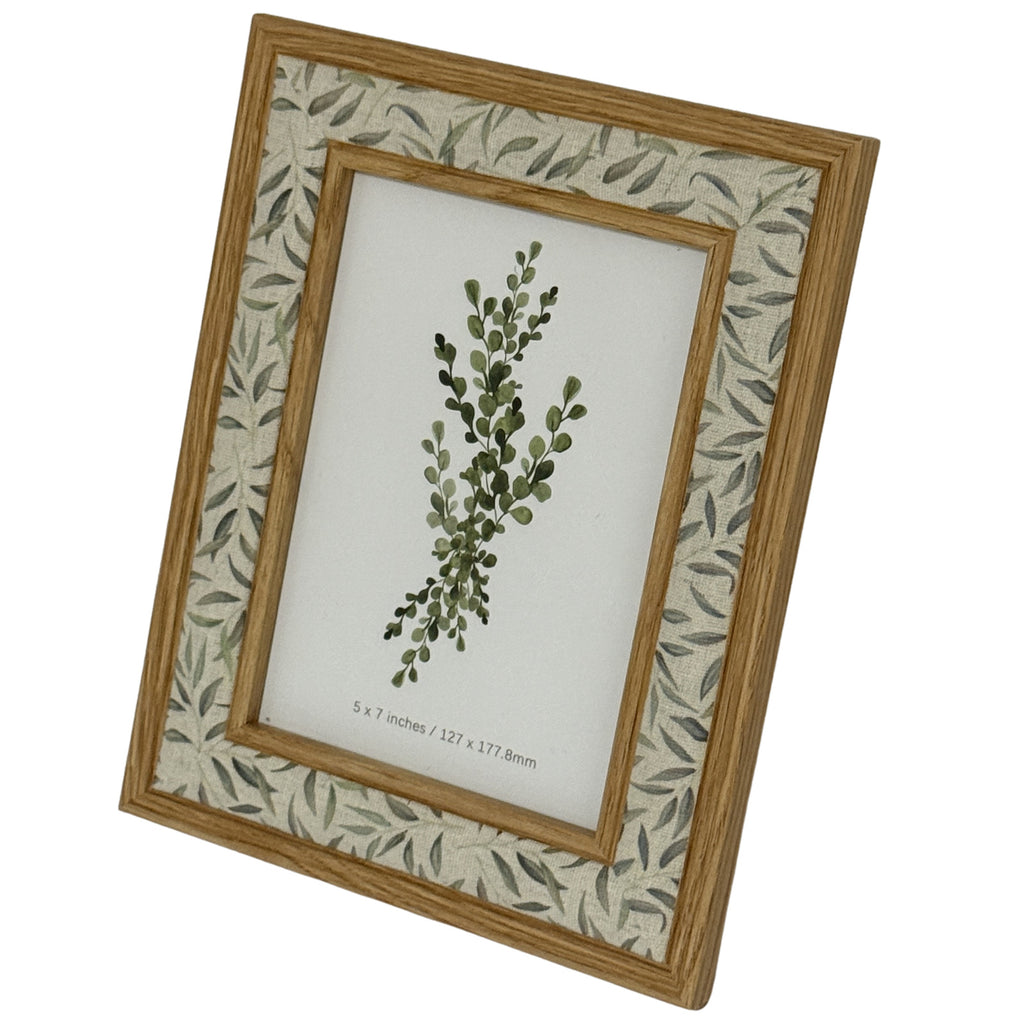 Angled view of the photo frame showing its double warm wood-tone trim around a pale band of scattered grey-green leaves.