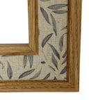 Close-up of a lower frame corner, showing grain-like markings on the warm wood-tone trim beside a pale textile-like band printed with grey-green leaves.