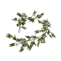 Artificial foliage garland curved against a white background, with mixed green leaves, small glowing light points and a visible battery box.