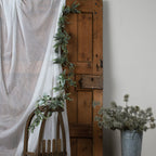 Foliage garland hanging beside white fabric against a wooden door and trailing over a wooden chair, with separate frosted greenery in a metal bucket nearby.