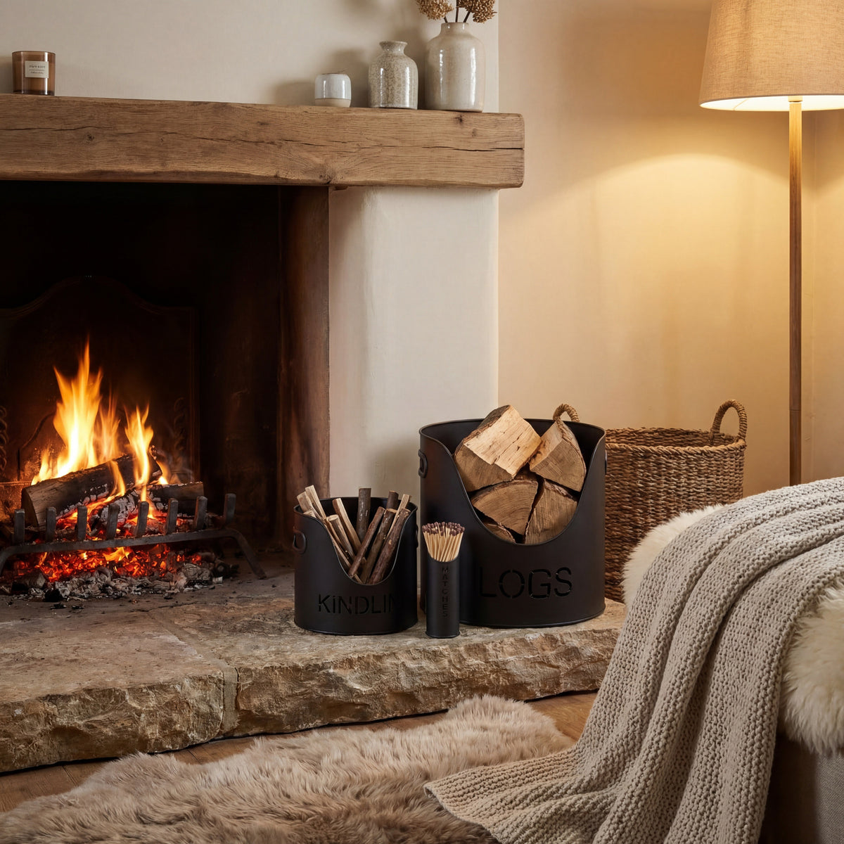 Lettered black buckets styled with logs and kindling beside a lit fireplace, with a matchstick holder, woven basket and knitted throw.