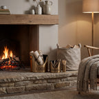Lettered bronze-toned buckets styled with logs and kindling beside a match holder on a stone hearth, with a lit fireplace, fabric log bag and fringed throw nearby.