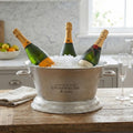 Lettered oval champagne cooler styled with ice and three bottles on a wooden table beside folded linen and a champagne flute.
