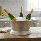 Lettered oval champagne cooler styled with ice and three bottles on a wooden table beside folded linen and a champagne flute.