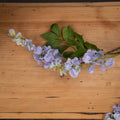 Lilac and creamy white artificial flower clusters with pointed green leaves laid across a wooden tabletop.