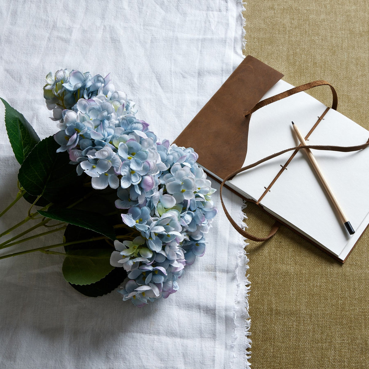 Pale blue and lilac artificial flower bouquet laid diagonally on white cloth beside an open brown-covered notebook and a pencil on a beige surface.