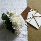 White artificial flower bouquet laid diagonally on white cloth beside an open brown-covered notebook and a pencil on a tan surface.