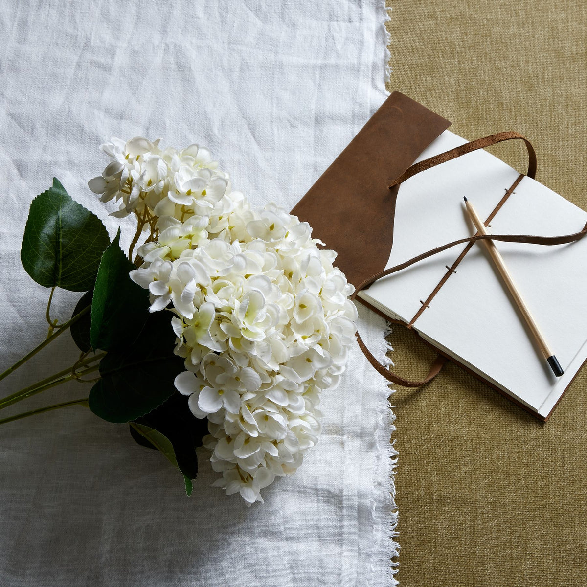 White artificial flower bouquet laid diagonally on white cloth beside an open brown-covered notebook and a pencil on a tan surface.
