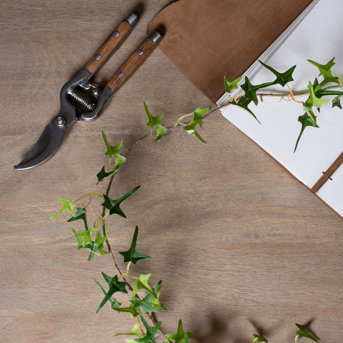 Artificial ivy garland curving across a wooden tabletop beside wood-handled secateurs and an open brown-covered notebook.
