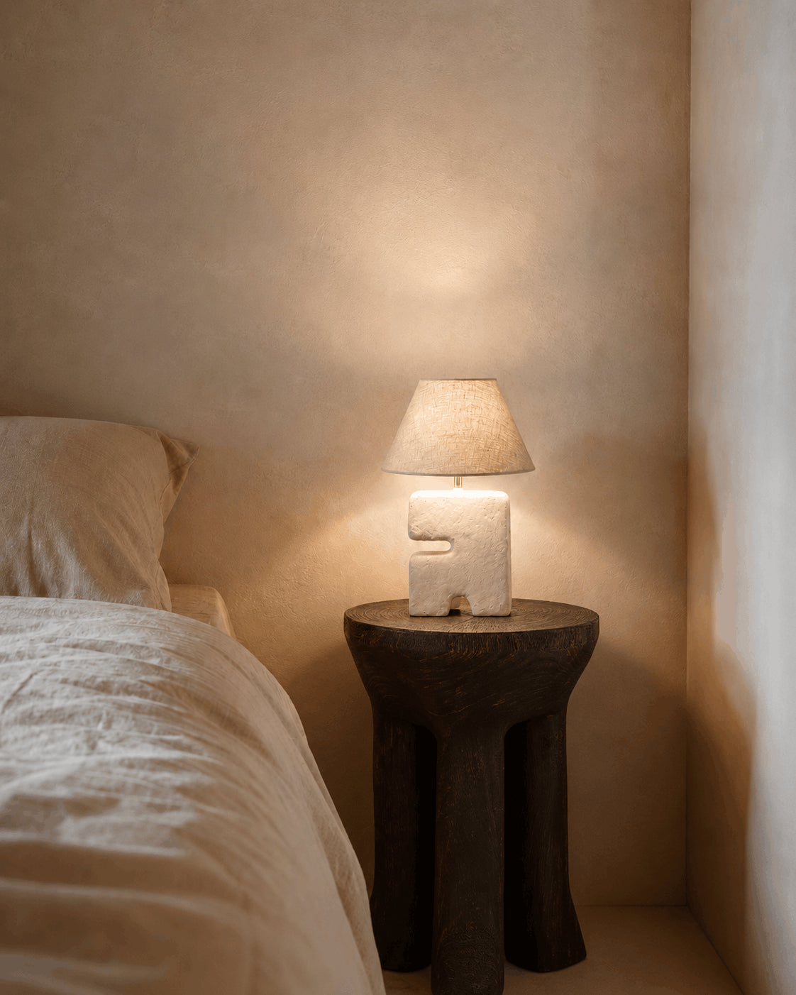 L’Objet Papier-Mâché Table Lamp – White, Small — lit on a dark side table