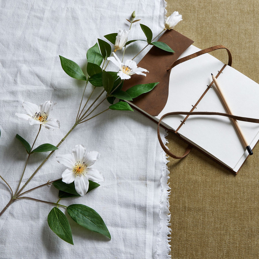 White artificial flowers and green leaves laid diagonally across pale cloth beside an open notebook and pencil on a tan textured surface.