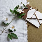 White artificial flowers and green leaves laid diagonally across pale cloth beside an open notebook and pencil on a tan textured surface.