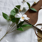 Close-up of a white artificial flower with a yellow and dark-tipped centre, pointed green leaves and closed buds on pale cloth beside a brown notebook cover.