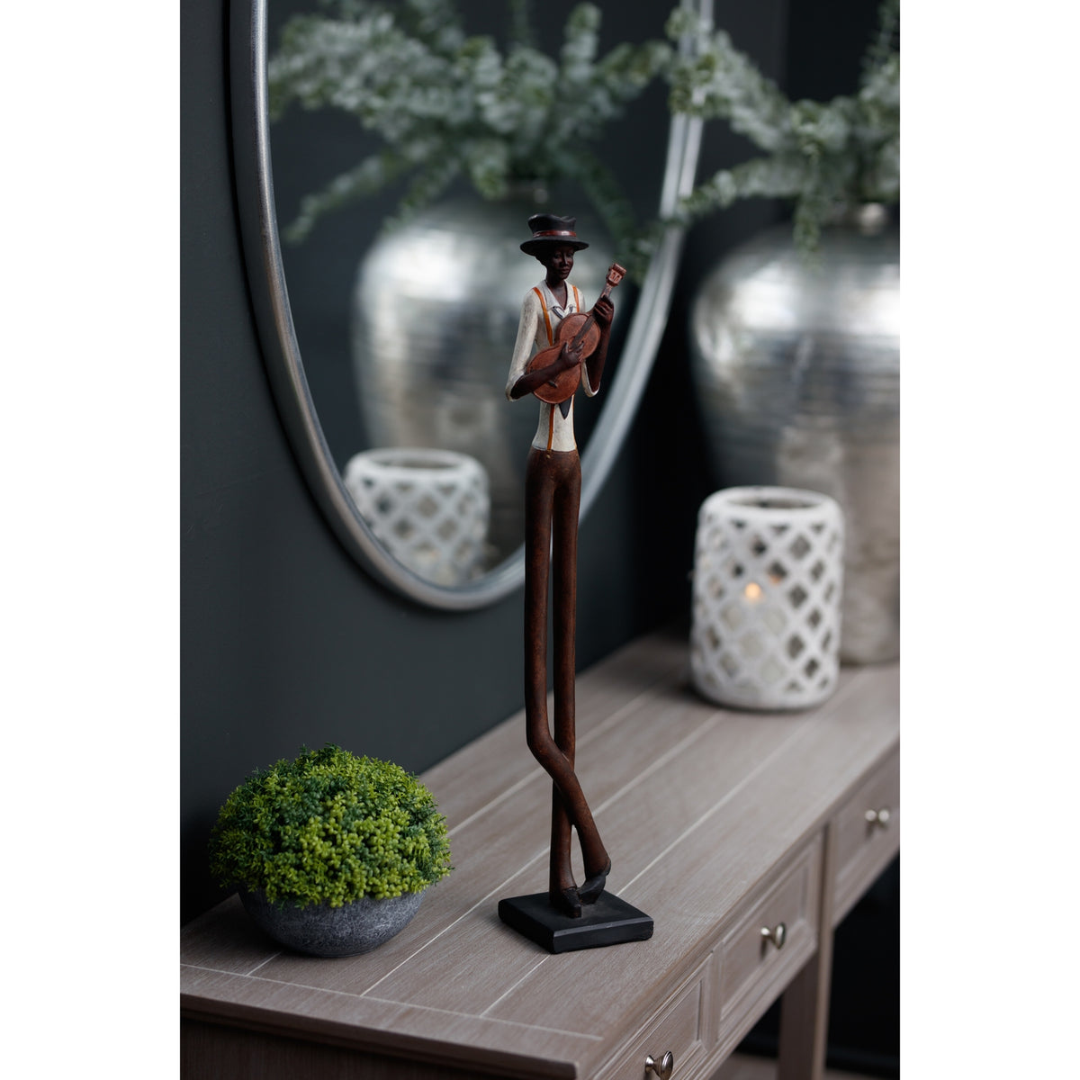 Full-length front three-quarter view of the guitarist figurine on a console, showing its crossed legs and dark base beside greenery and a white lantern.