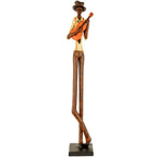 Full-length musician figurine against white, with elongated brown legs crossed near the ankles, a cream shirt, orange braces and a guitar on a dark rectangular plinth.