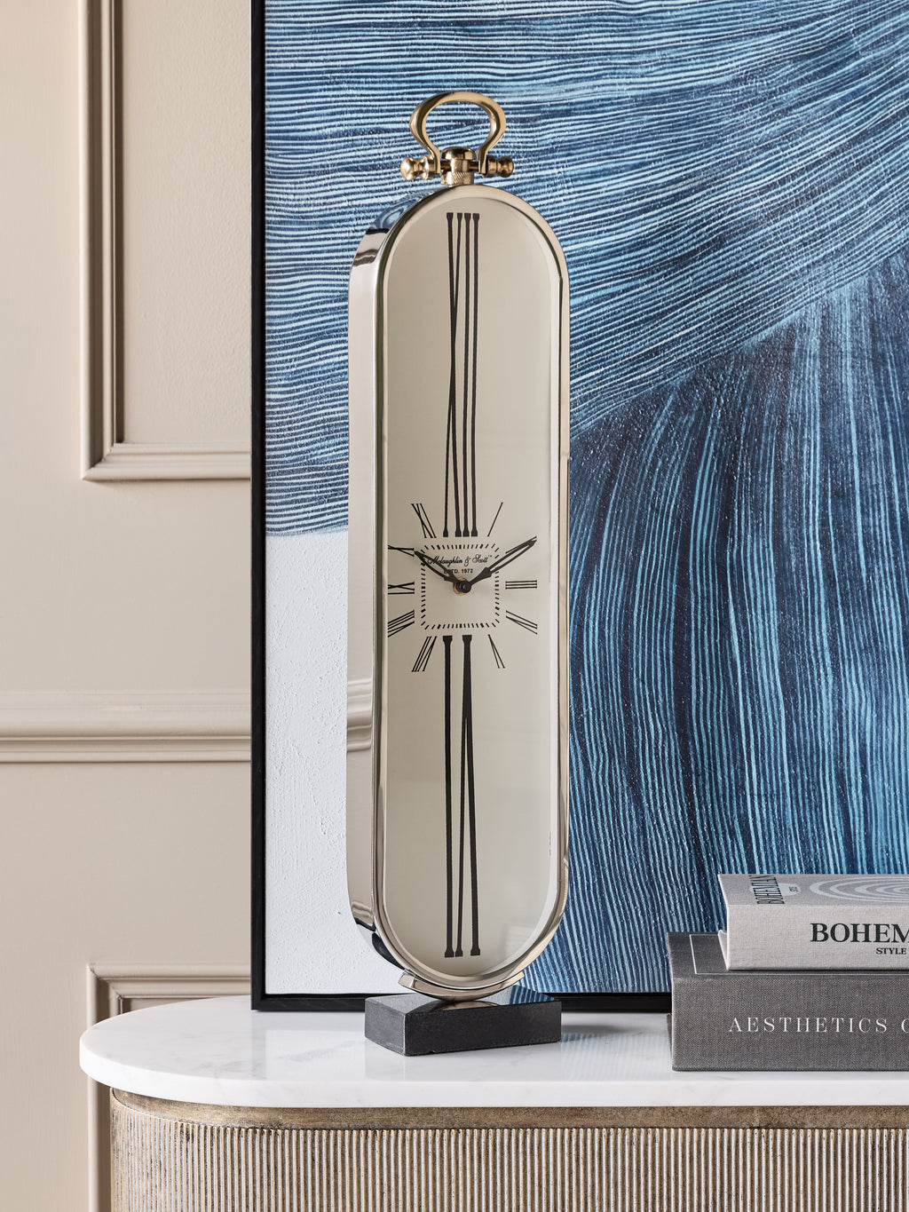 Oval clock displayed on a white-topped console beside stacked books, with a large blue abstract artwork behind it.