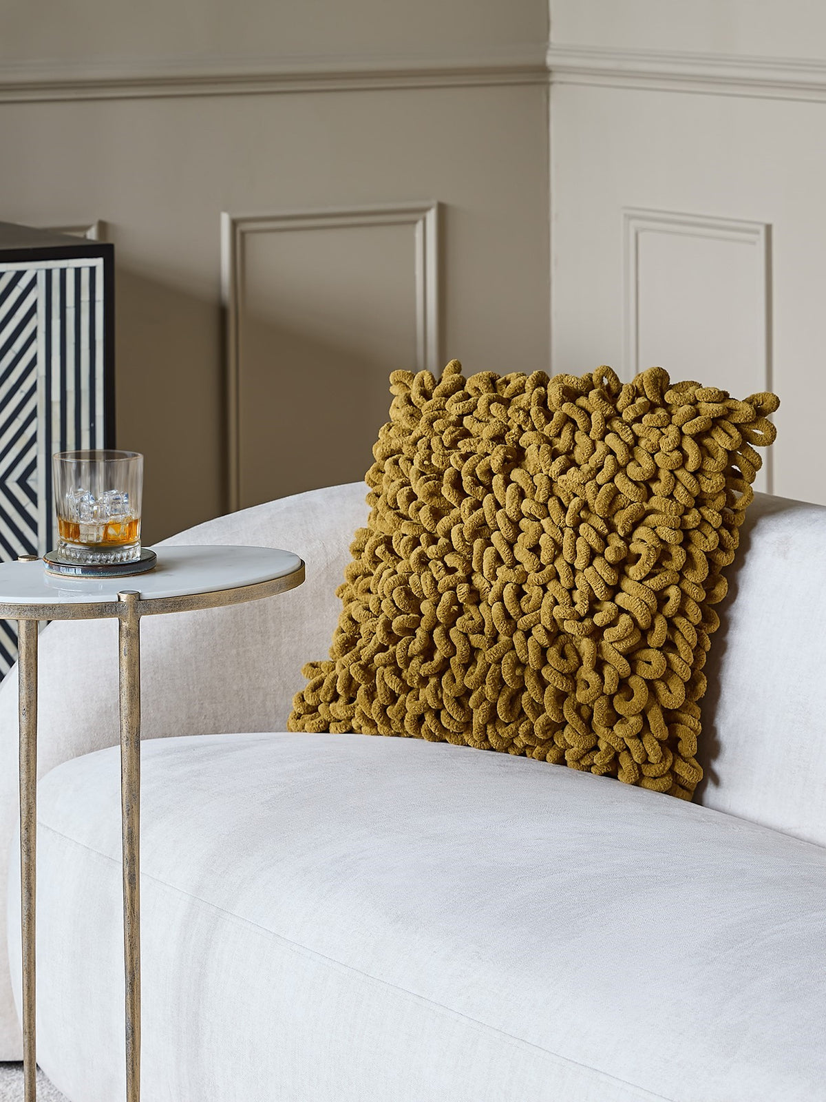 Mustard looped cushion on a cream armchair beside a round side table, with a panelled wall behind.