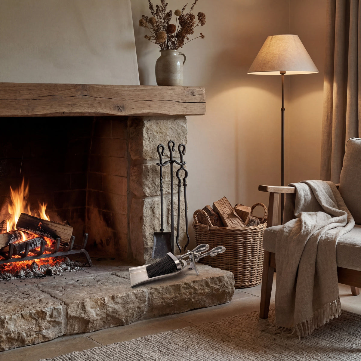 Looped scoop and brush arranged on a stone hearth beside a lit fireplace, with upright fireplace tools, a woven log basket and an armchair nearby.
