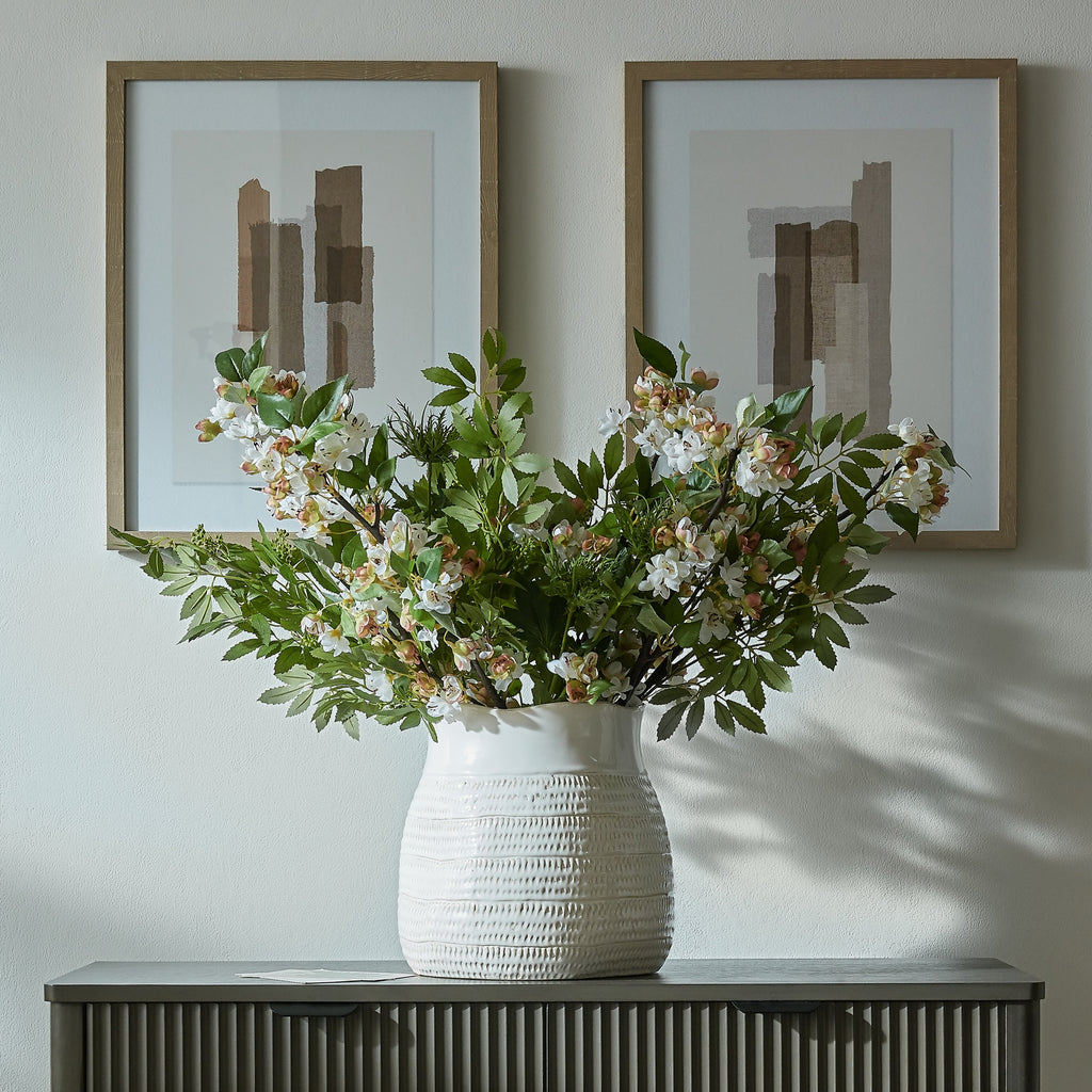 White patterned vase holding a spreading arrangement of green foliage and pale flowers on a dark sideboard beneath two abstract prints.