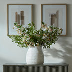 White patterned vase holding a spreading arrangement of green foliage and pale flowers on a dark sideboard beneath two abstract prints.