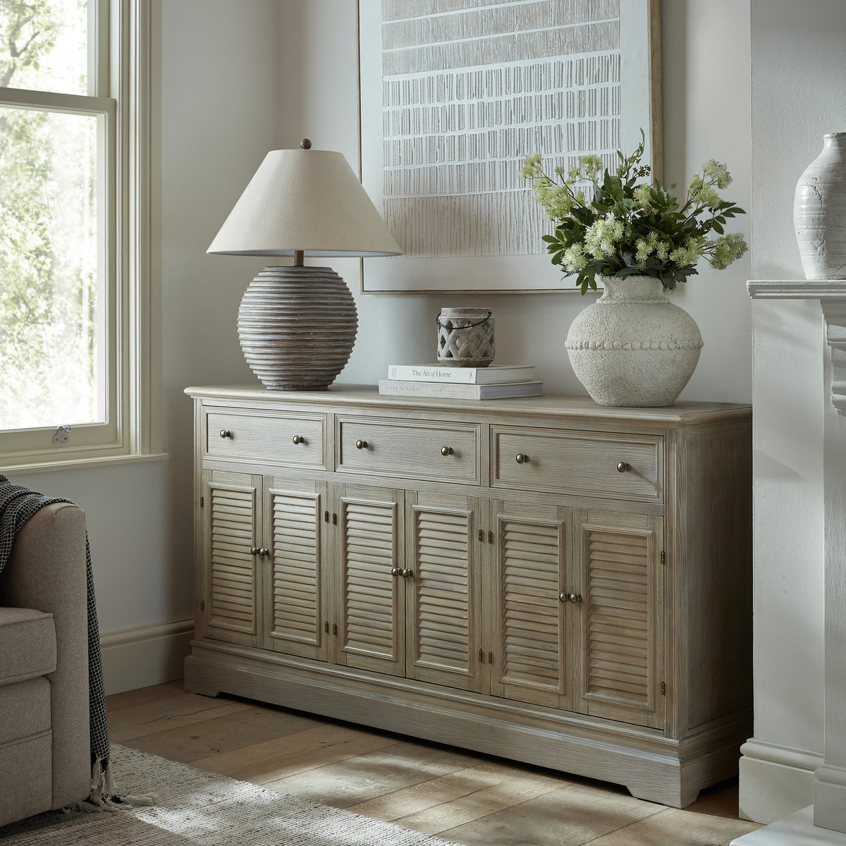 Dresser base beside a window and fireplace, styled with a table lamp, books and a vase of flowers beneath a large framed artwork.