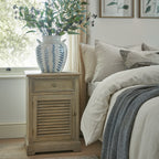 Cabinet beside a cream-dressed bed and window, styled with a blue-and-white vase of leafy branches beneath botanical prints.