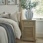Cabinet beside a bed with cream bedding, styled beneath a window with a blue-and-white vase of leafy stems on top.