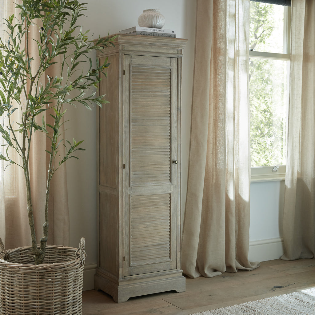 Louvred cabinet beside a window with pale curtains, styled with books and a pale vase on top and a leafy tree in a woven basket alongside.